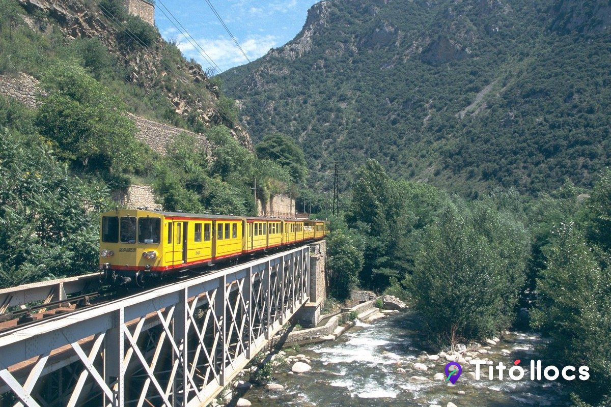 El Tren Groc passant per un pont de ferro sobre el riu, entre les muntanyes del Conflent